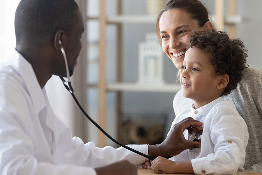 A healthcare professional uses a stethoscope to listen to a young child’s chest while a caregiver sits beside the child in a clinical setting