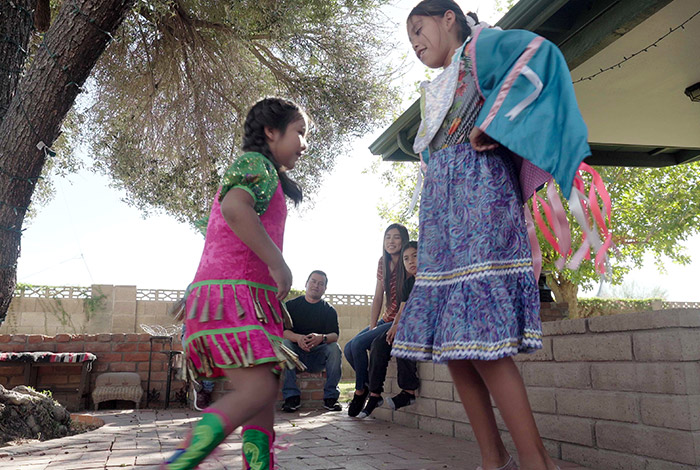 Two girls dancing in traditional Native American apparel while their family watches. 