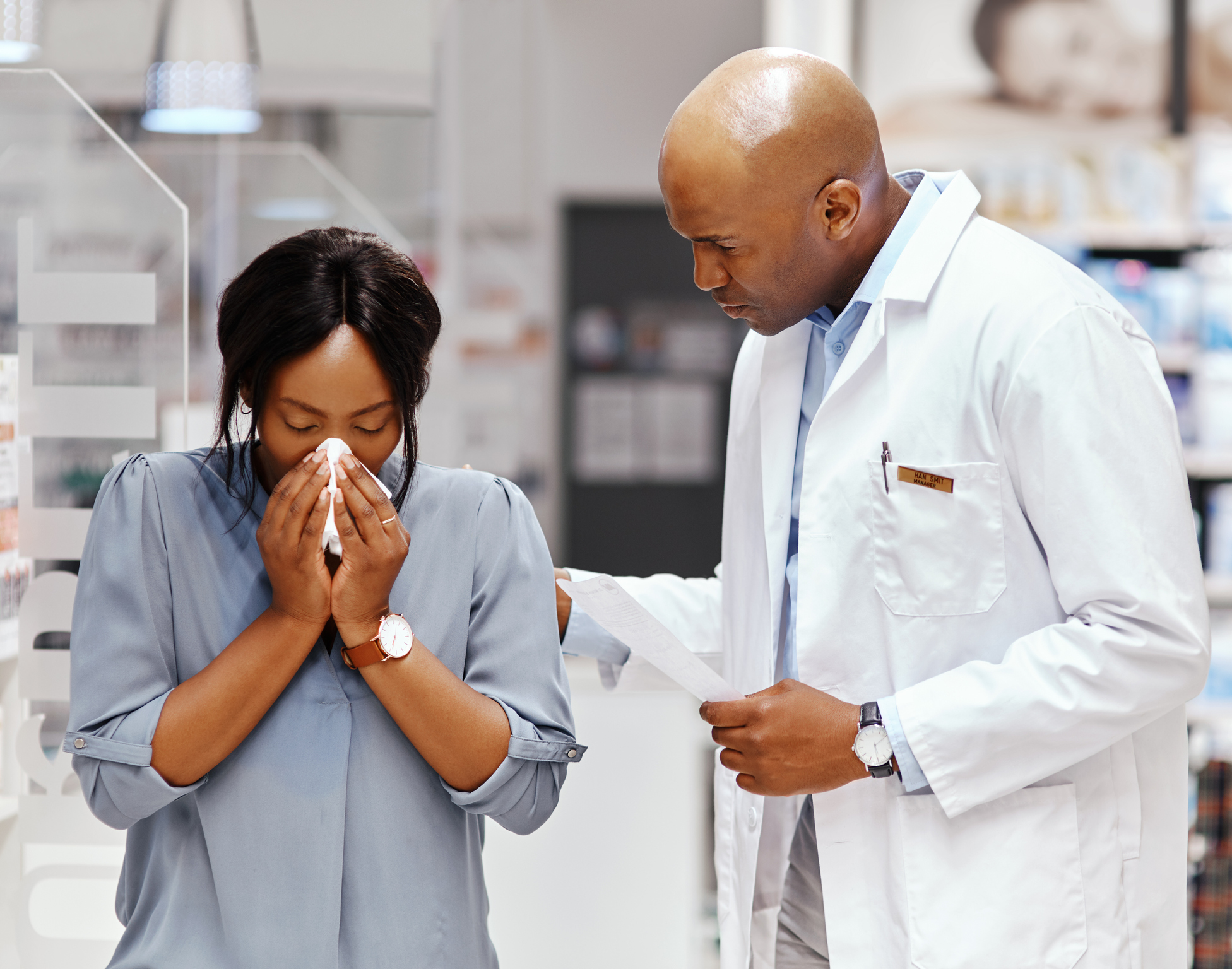 pharmacist looks on as woman uses tissue to relieve flu or coronavirus symptoms