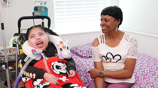 Adult sitting beside a child in a wheelchair on a bed, with medical equipment.