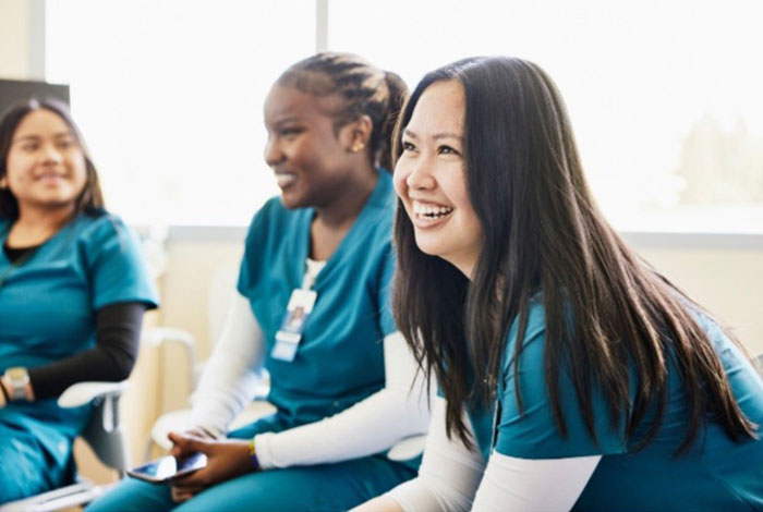 Three people wearing teal medical scrubs sit together indoors, engaged in a group discussion.