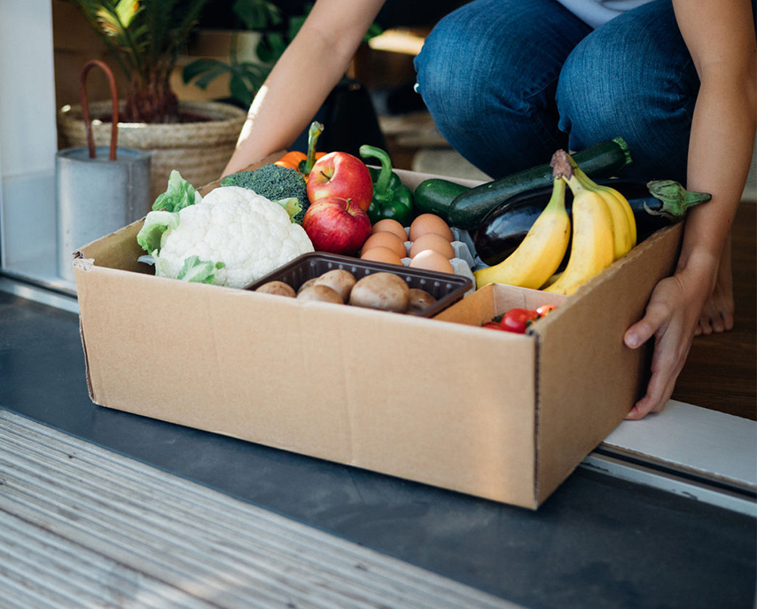 A person kneeling at a doorway holding a cardboard box filled with fresh groceries